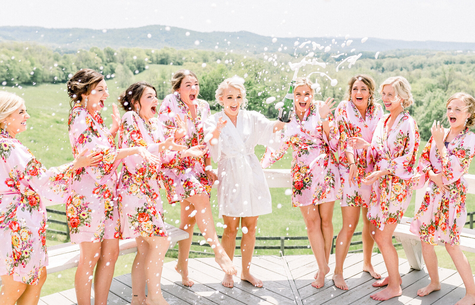 A bride in a white robe surrounded by bridesmaids in pink floral robes as champagne sprays over them on an outdoor deck