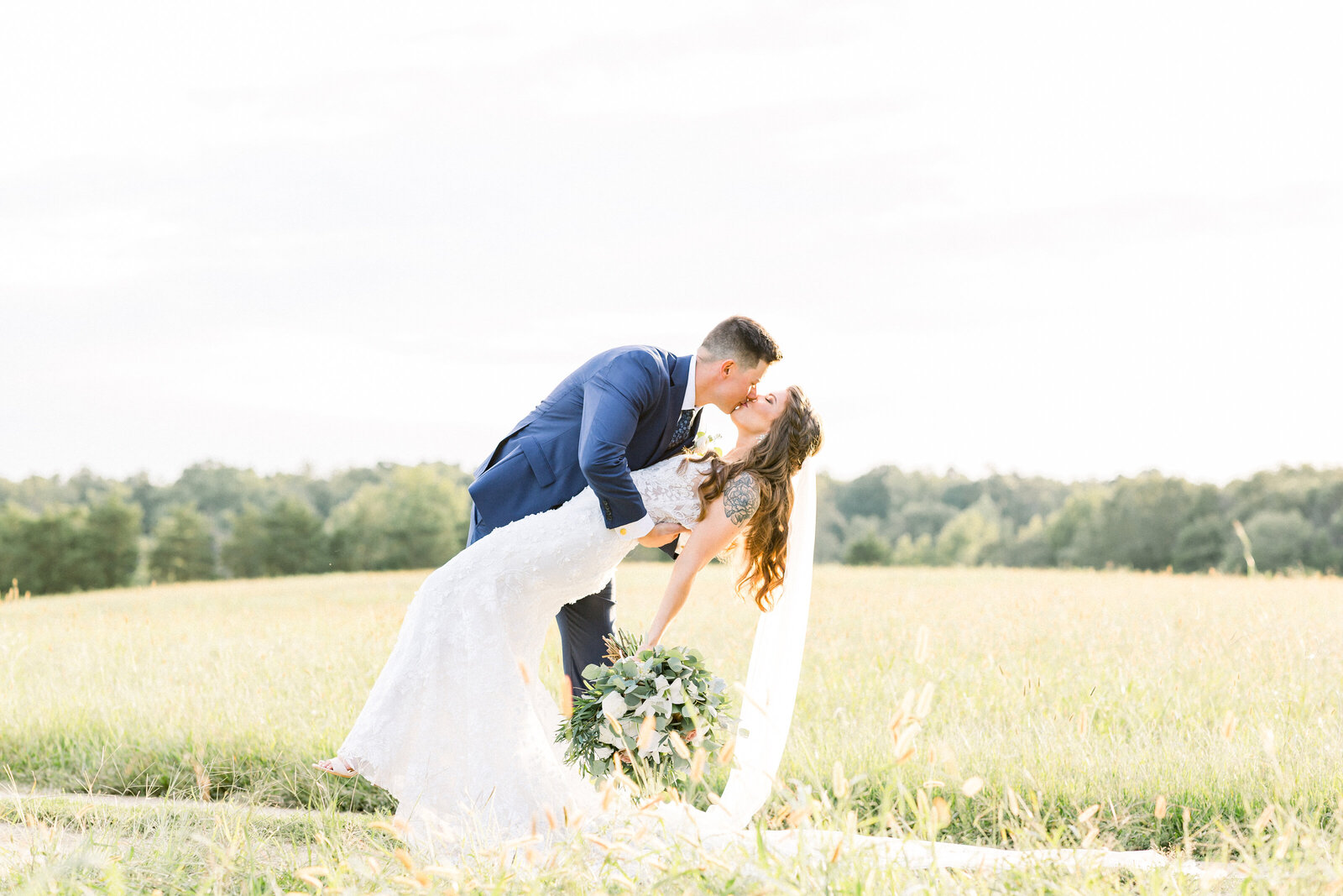 A groom in a navy suit dipping his bride for a kiss in an open field of tall grass at golden hour